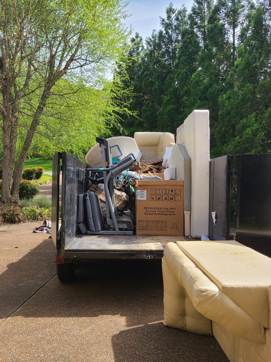 Trailer loaded with a couch, mattress and boxes in a wooded driveway