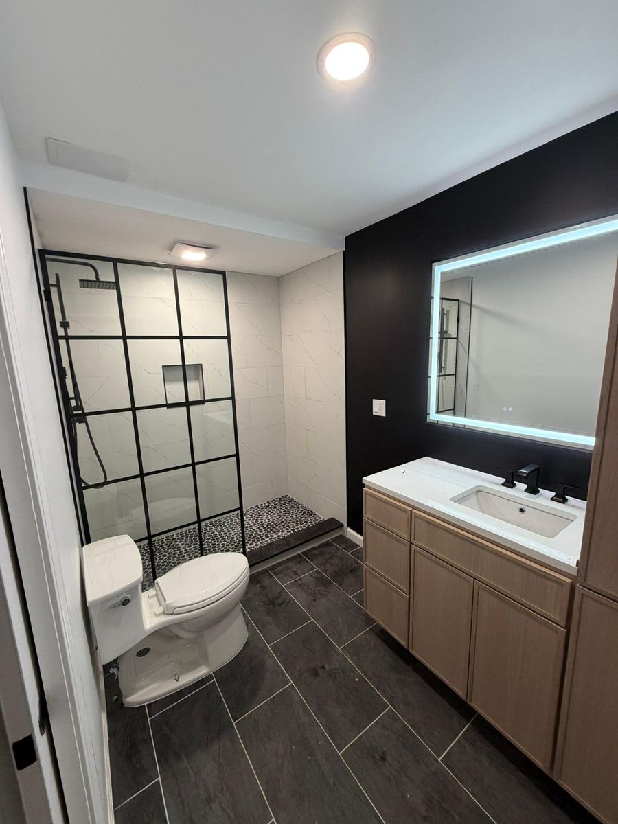 Remodeled bathroom with black-framed glass shower, dark tile floor, and lit vanity mirror
