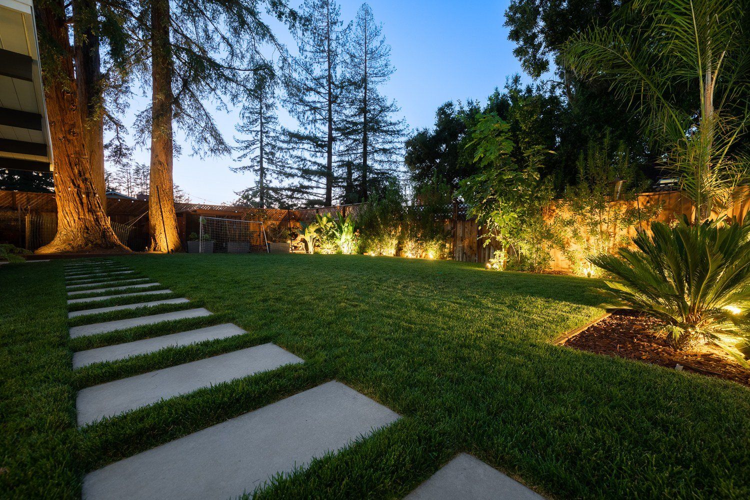 Long pale concrete pavers stepping across a green lawn at dusk, with uplit tall trunks, large-leaf plants and a palm glowing along a wood fence.