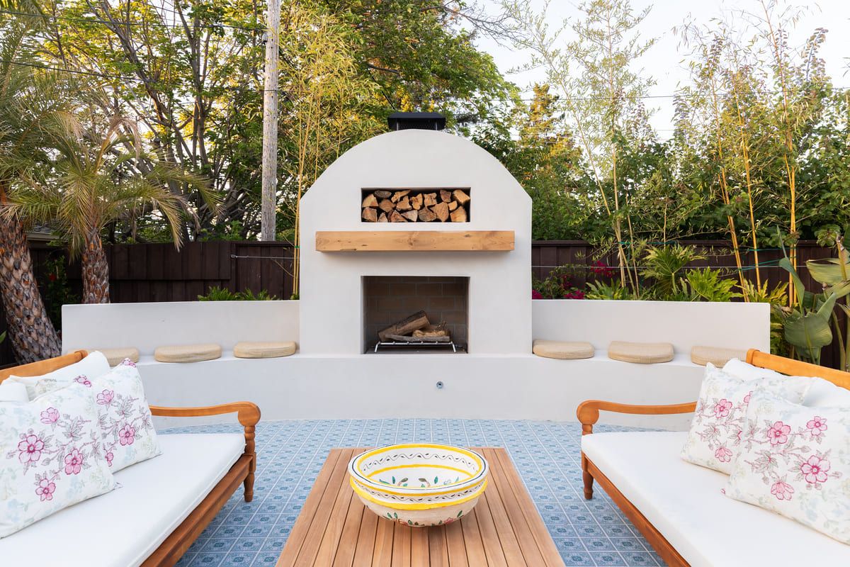An arched rendered outdoor fireplace with a timber mantel and log store, cushioned seat wall facing it