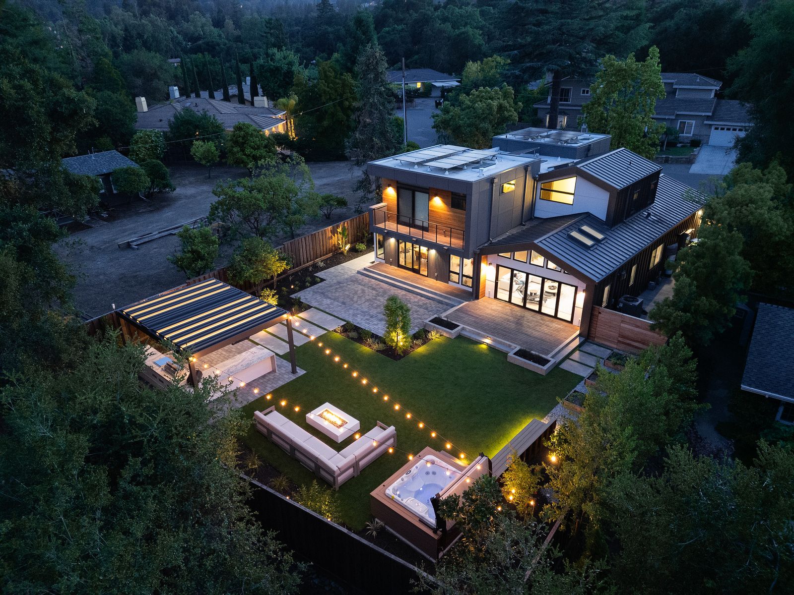 Twilight aerial of the whole garden — lawn, lit terrace, fire table and pergola, with the modern house behind