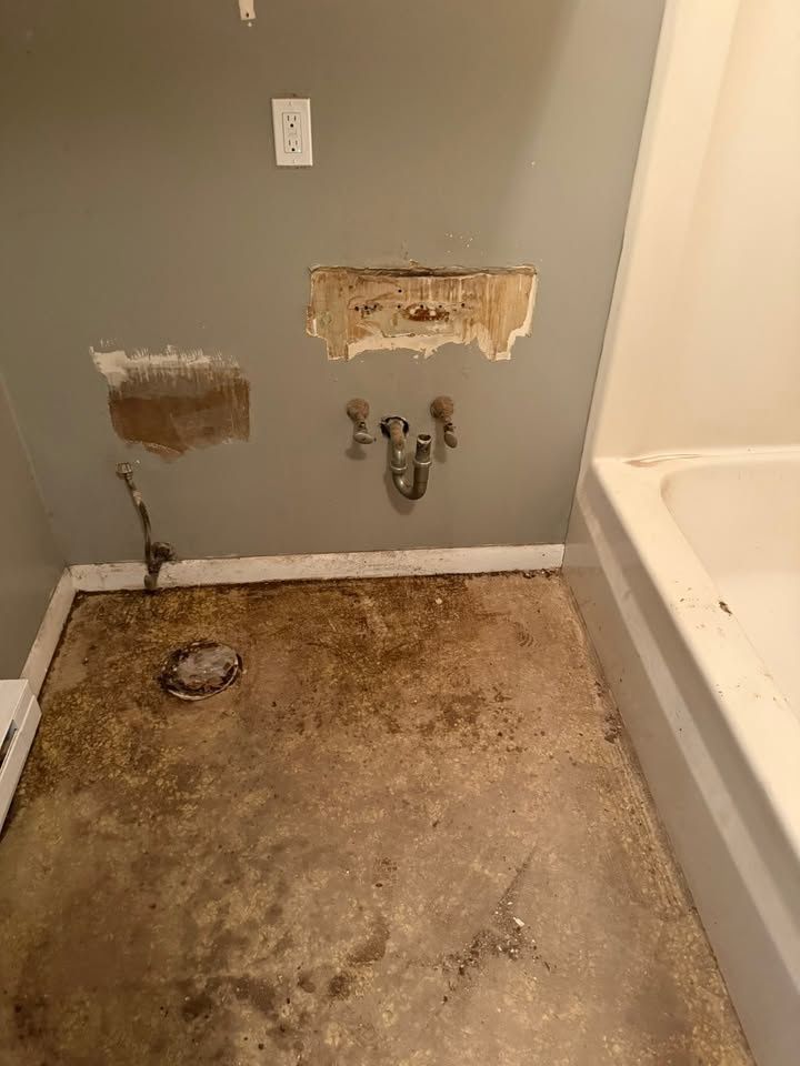 Bathroom wall with the sink removed, the supply lines left in place and patches cut out of the sheetrock above a stained floor