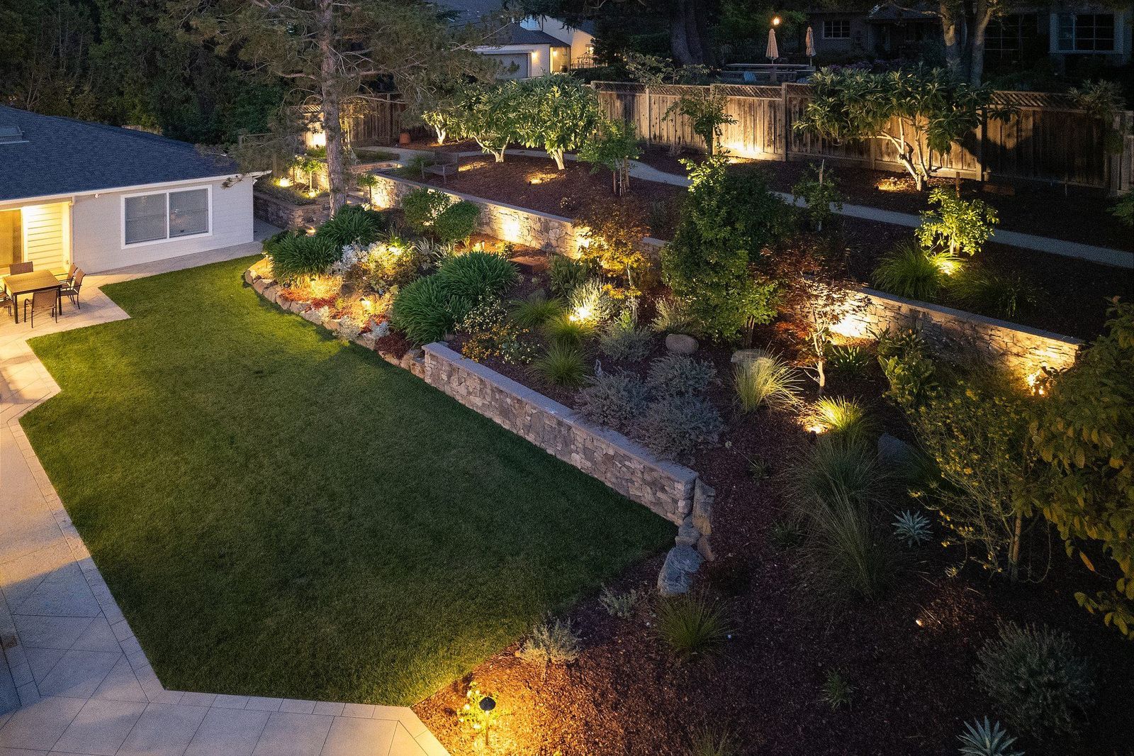 Twilight aerial of a graded hillside — lawn, planted terraces and mature trees under low-voltage lighting