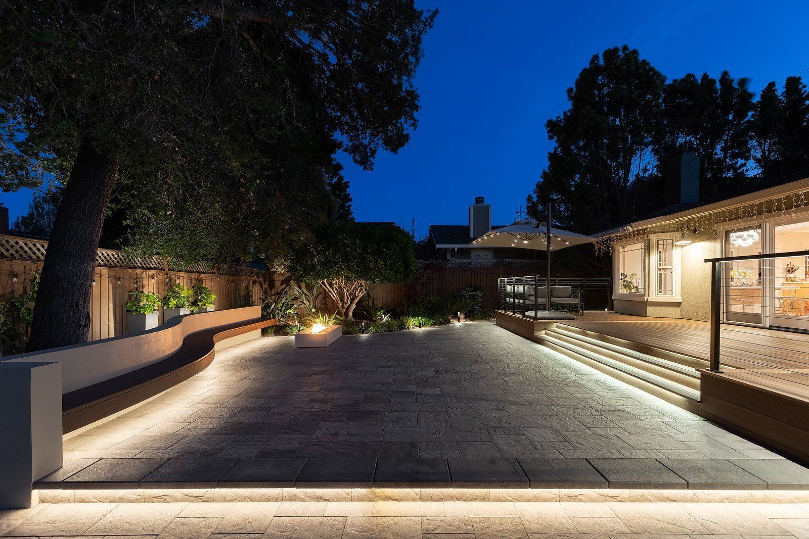 Twilight view across a gray paver court with a curved bench glowing with under-lighting, a fire table, and a stepped wood-look deck beside the house