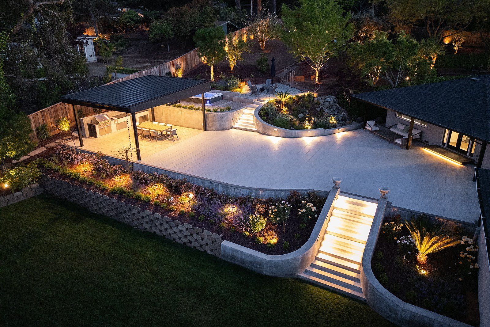 Twilight aerial of curved concrete terraces, a lit central stair, a dark-roofed pavilion with an outdoor kitchen and planted beds glowing with garden lighting