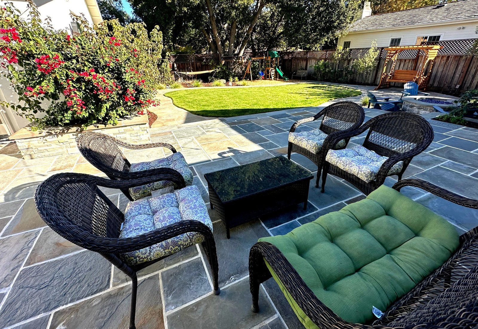 Bluestone patio with wicker armchairs and a green cushioned bench, a curved lawn beyond, a flowering bougainvillea in a stone planter and a fire pit area at the right.