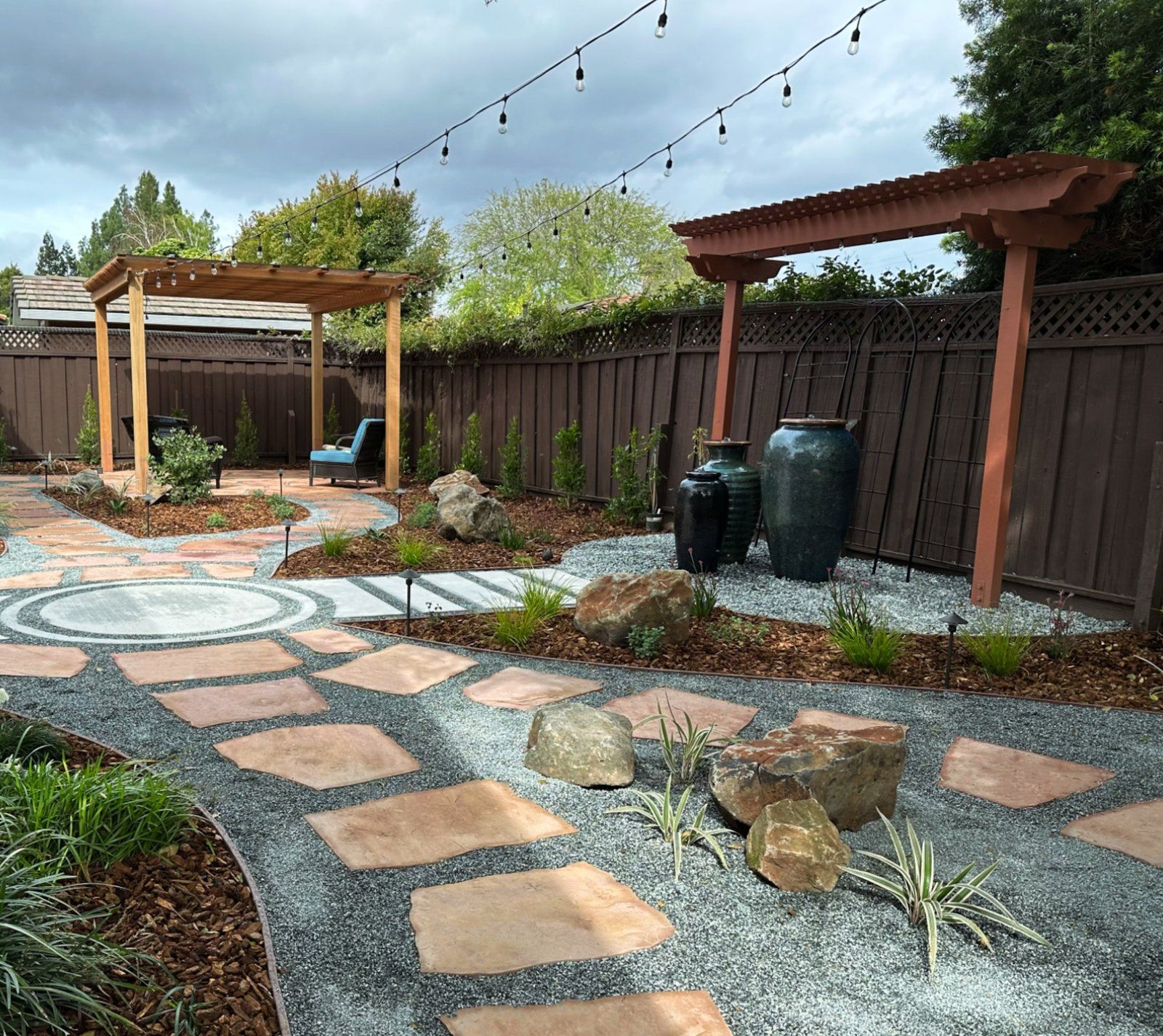 Terracotta flagstone stepping stones set in pale gravel past boulders and grasses, with two wood pergolas, large teal ceramic jars and string lights across the yard.