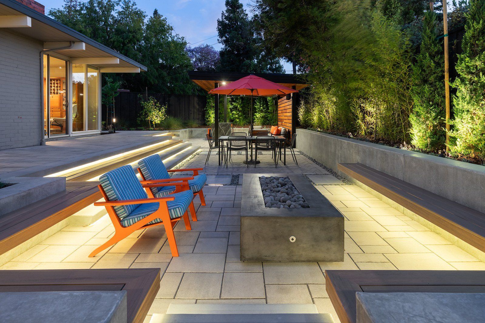 Twilight view of a paver terrace with a concrete fire table, striped lounge chairs, lit steps and a wood-roofed pavilion with an orange umbrella