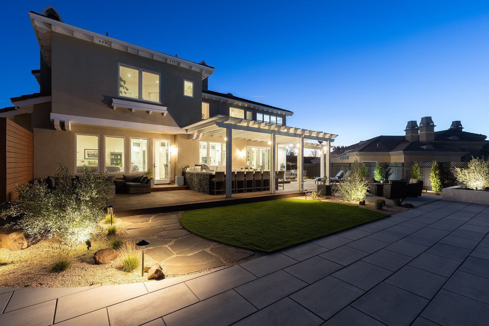 Twilight view of the back of the house, a white pergola over a deck and kitchen island, with a curved lawn, flagstone path and lit gravel beds in the foreground
