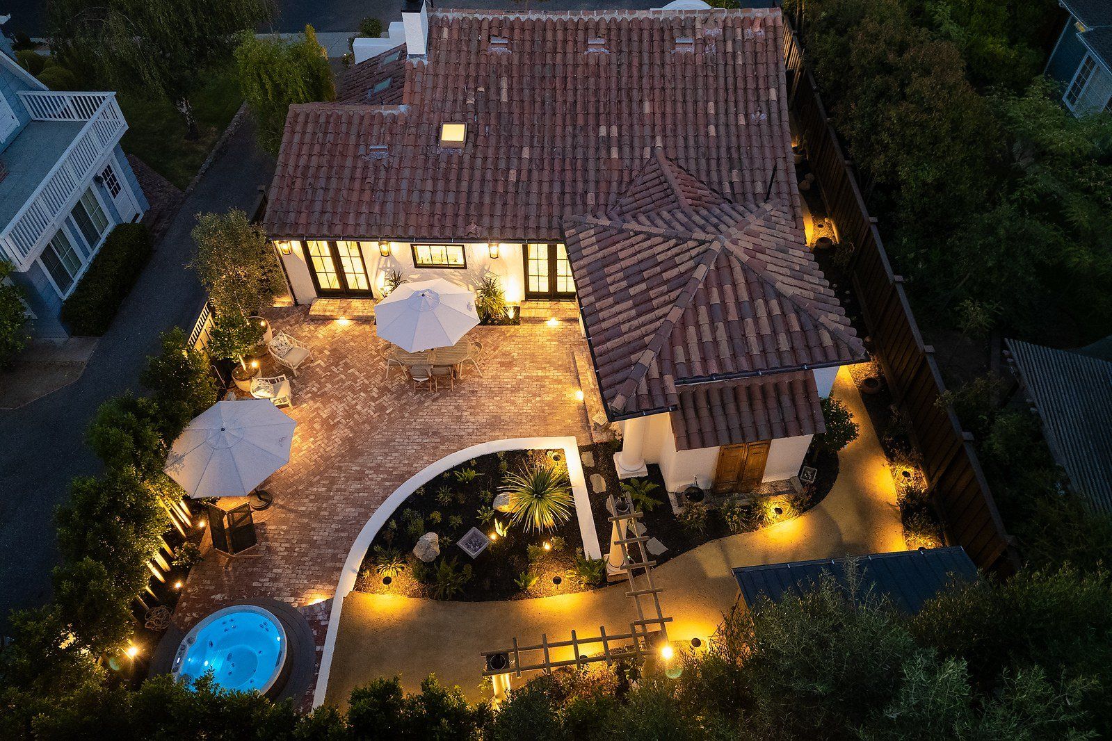 Twilight aerial of a herringbone brick courtyard with two umbrellas, a curved stucco planter bed, a decomposed granite path and warm landscape lighting beside a tile-roofed house