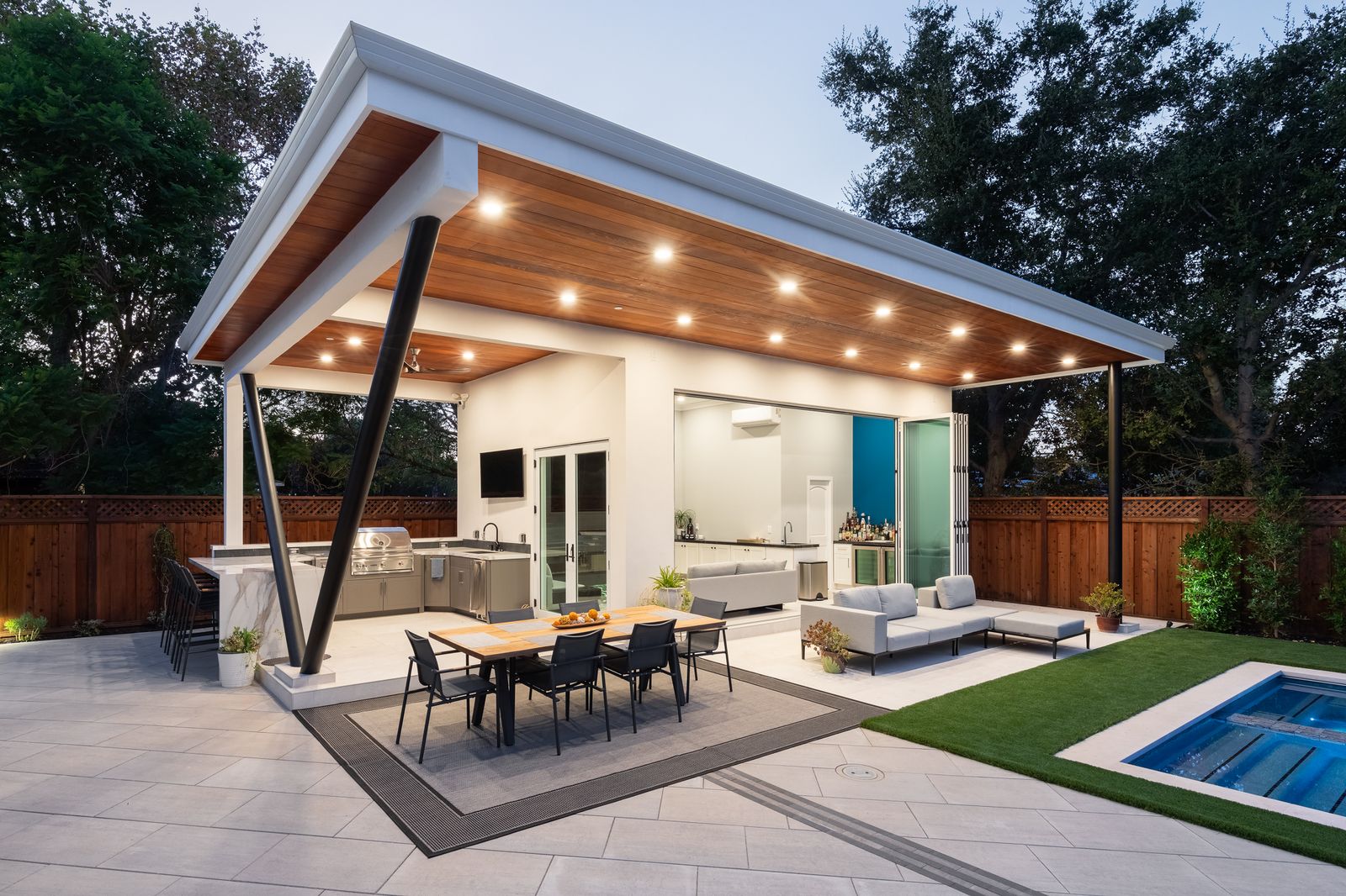 The cabana lit at dusk — timber-lined ceiling on an exposed steel strut, outdoor kitchen and lounge, pool beyond