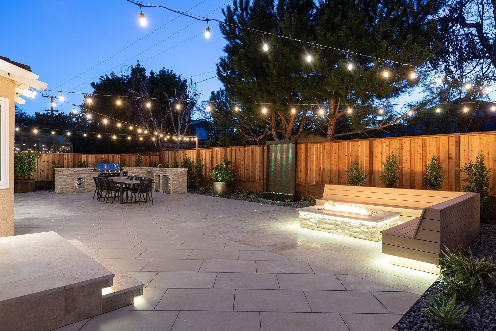 Twilight view across the pale stone patio, with a lit stacked-stone fire bench at right, a water wall in the fence and the outdoor kitchen and dining table at left under string lights