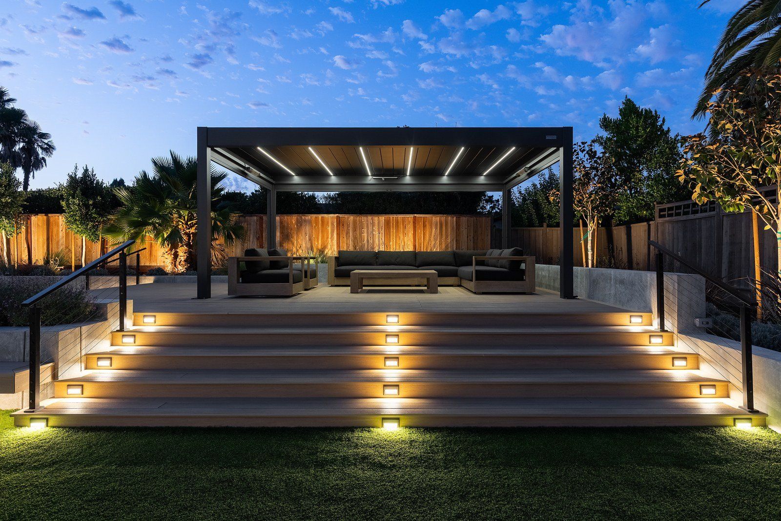 Twilight view of a raised deck with five wide lit steps, cable railings and a louvered pergola lit with white strip lights over a dark sofa lounge