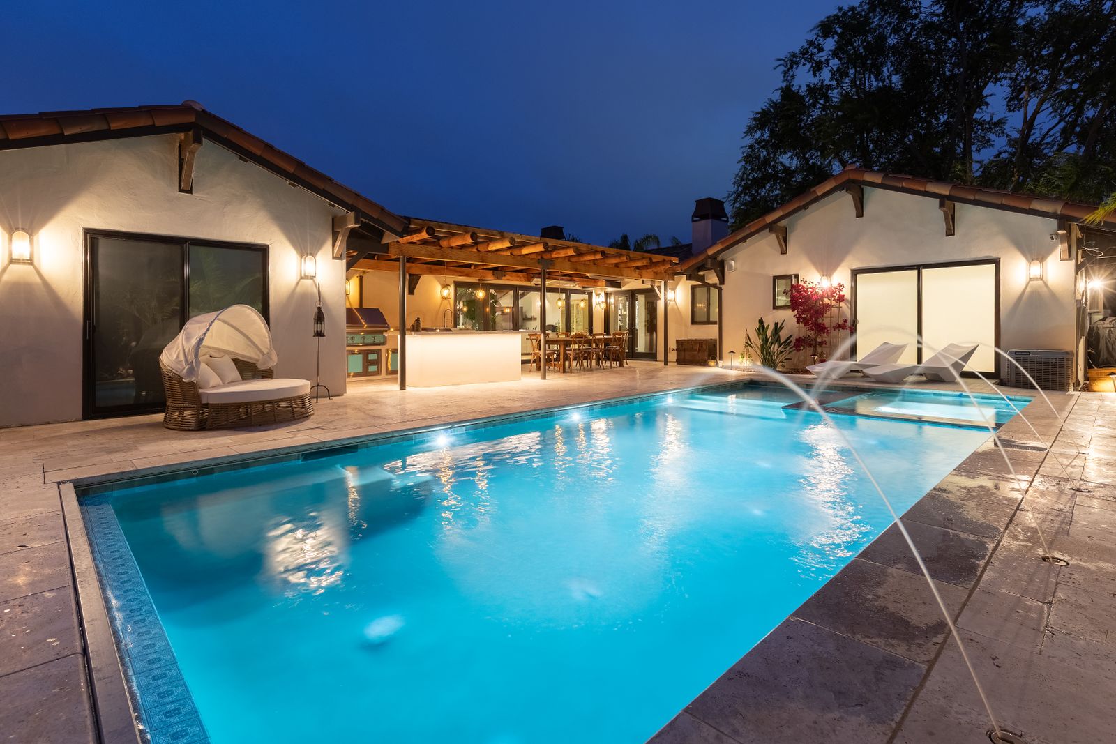 The pool at dusk with deck jets arcing over it, the Spanish-style house and its covered terrace lit behind