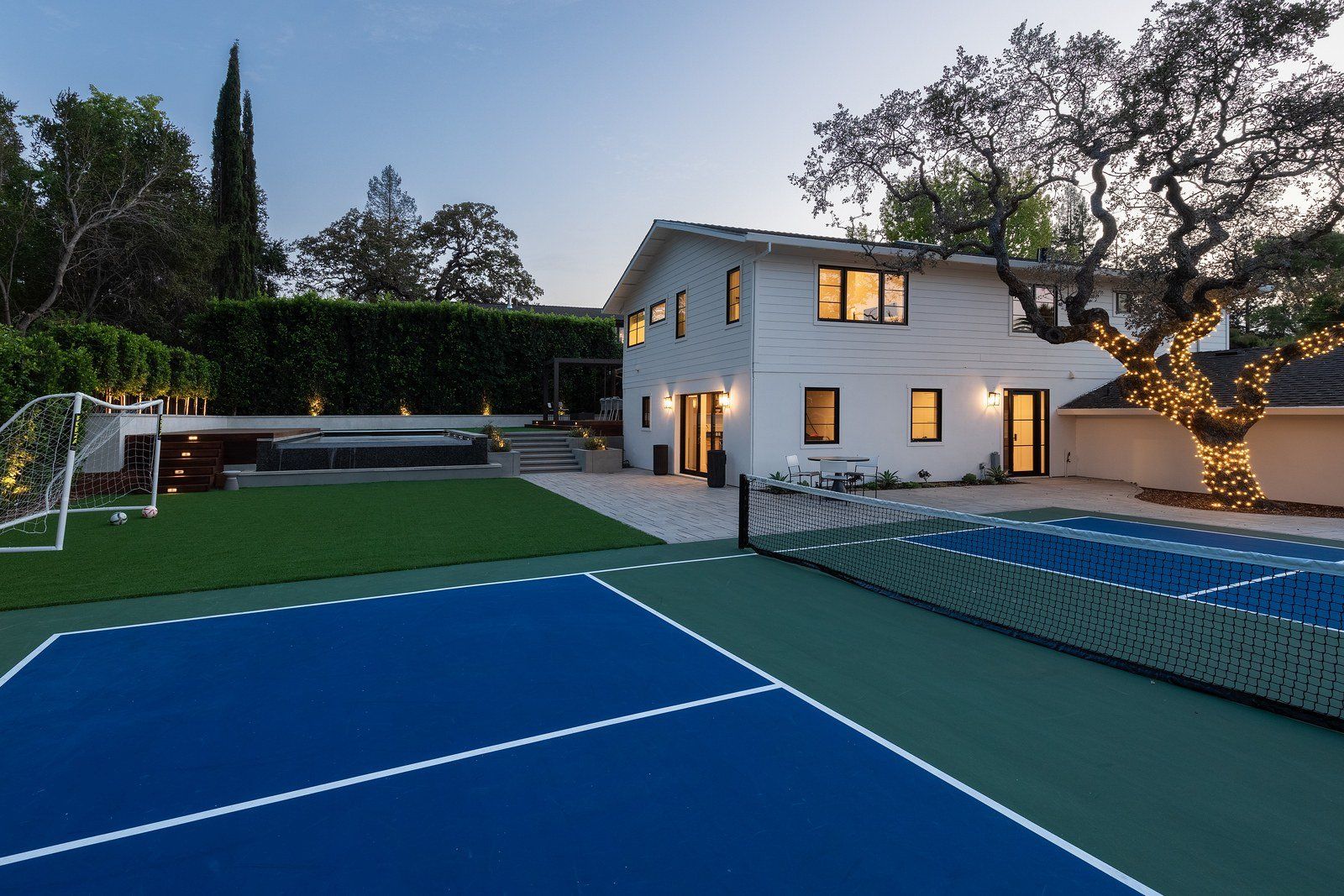 Twilight view across a paver patio and lawn to a white modern farmhouse with black windows, a lit oak tree wrapped in string lights and a stepped wall at the far end