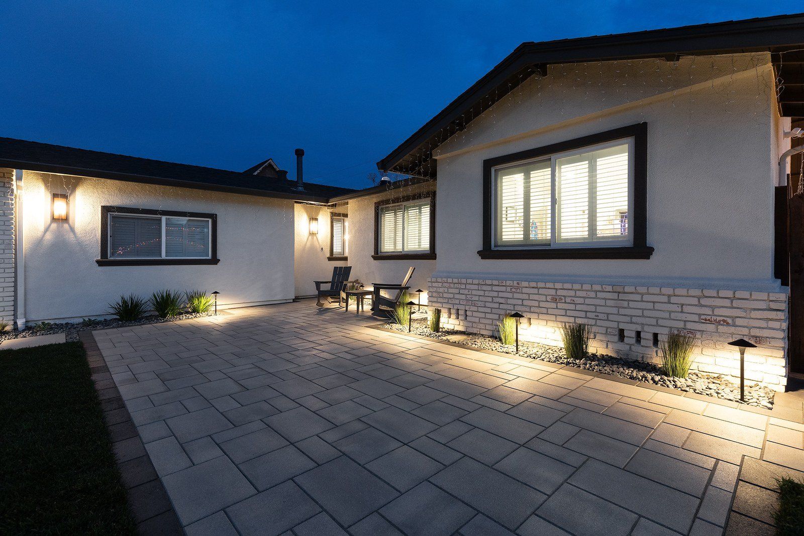 Twilight view across a gray paver front court, with a pair of Adirondack chairs by the entry and warm path lights washing a whitewashed brick base