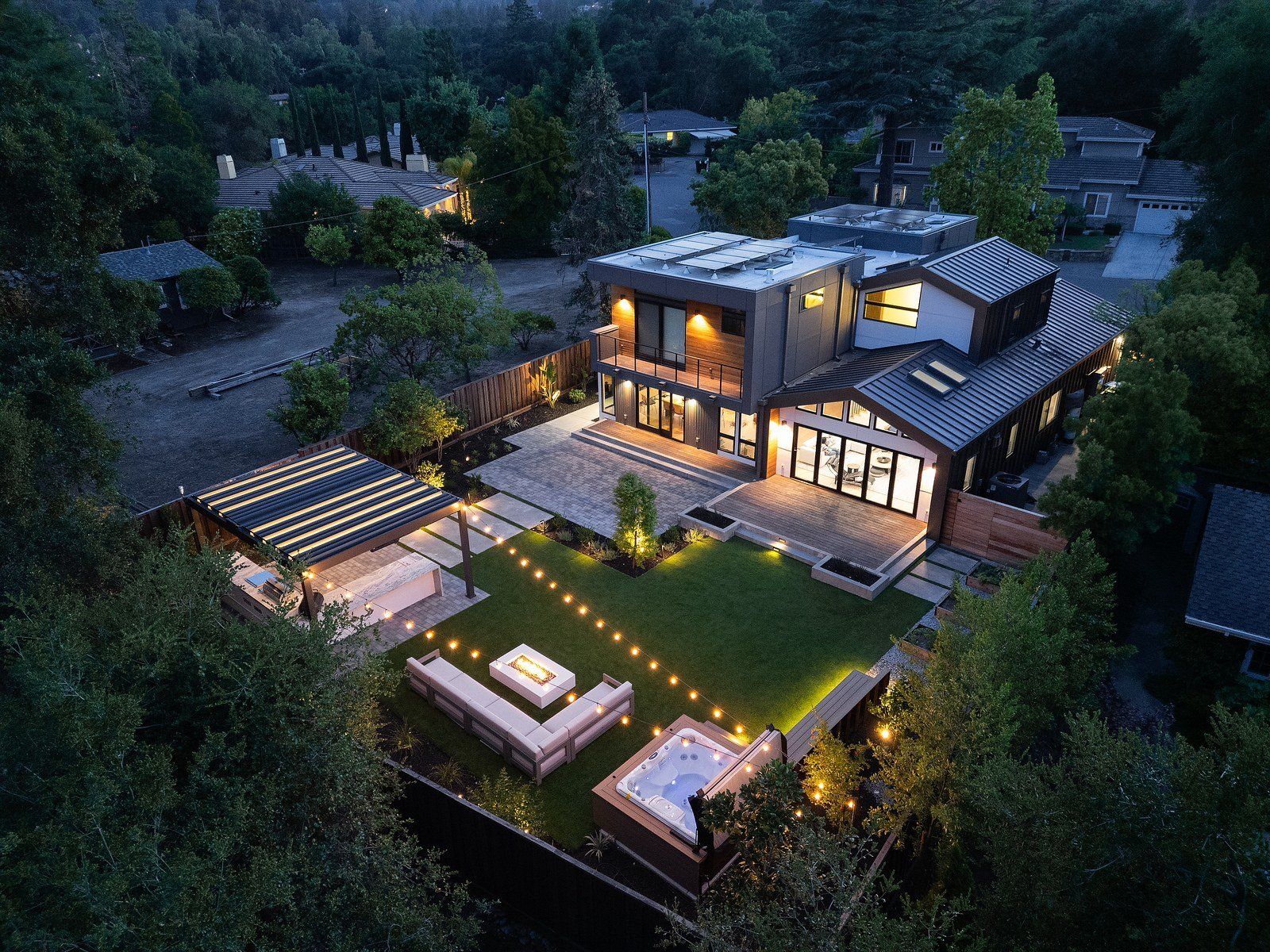 Twilight aerial of a modern house and its backyard, a lit lawn with a white fire table and sofa, a raised deck and a louvered pergola kitchen