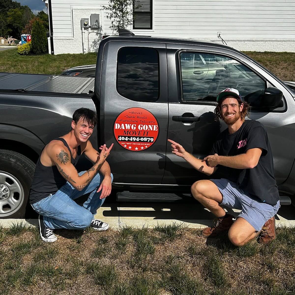 Two men crouching beside a gray pickup truck with a round Dawg Gone Move It sign on the door