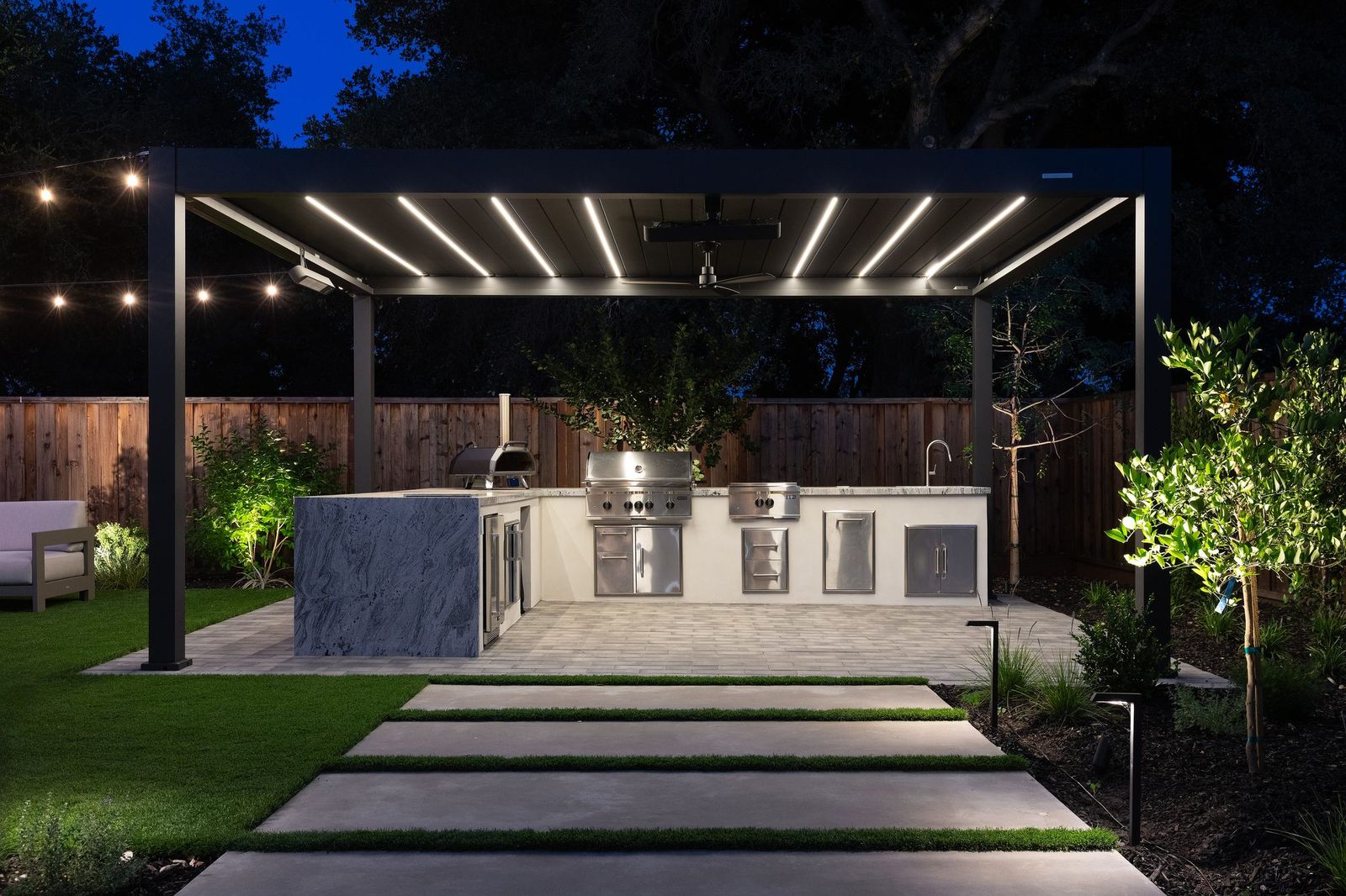 Outdoor kitchen lit at night under a louvered pergola, built-in appliances and stone worktops