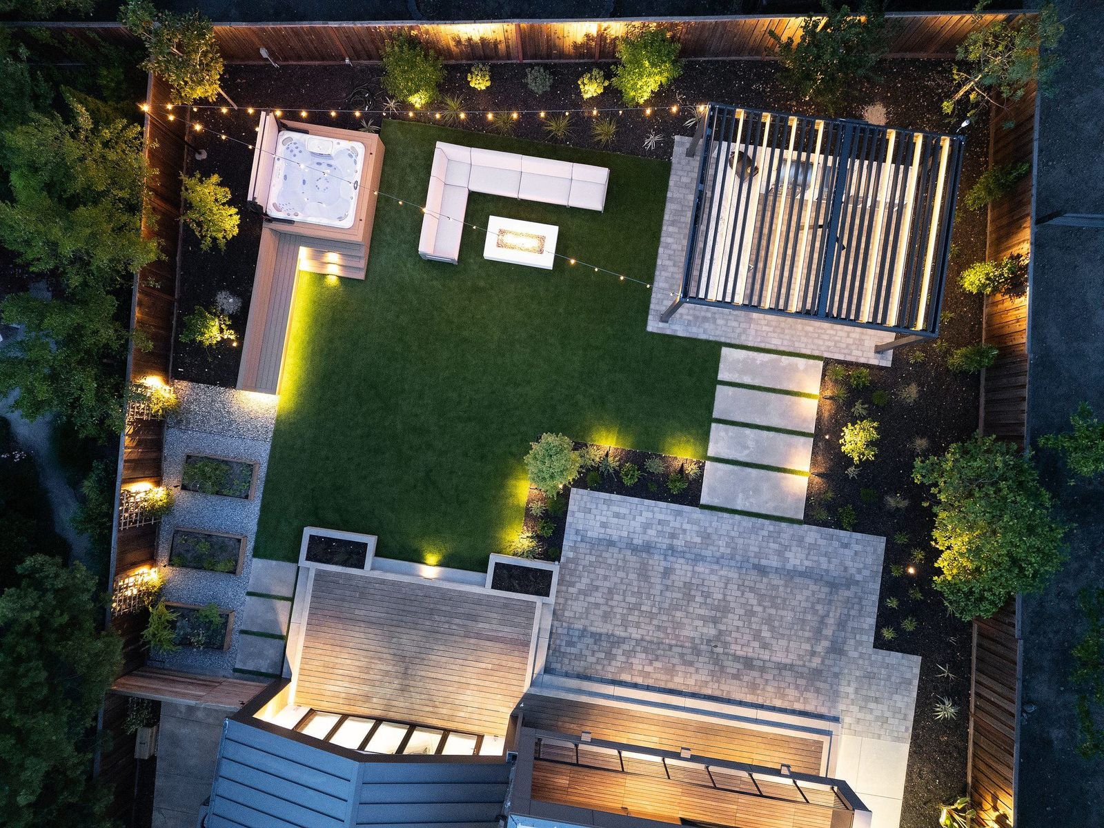Overhead aerial at night of a completed design — lit paving, fire table, seating and planted borders read as a plan