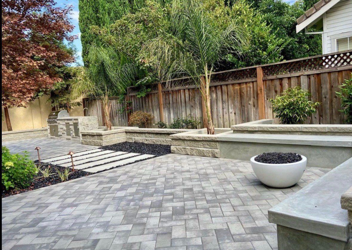 Gray herringbone paver patio with pale stepping-stone slabs set in black pebbles, copper path lights, a stone-faced outdoor kitchen at the left, stacked-block planter walls with palms, and a round white fire bowl on a capped seat wall at the right.