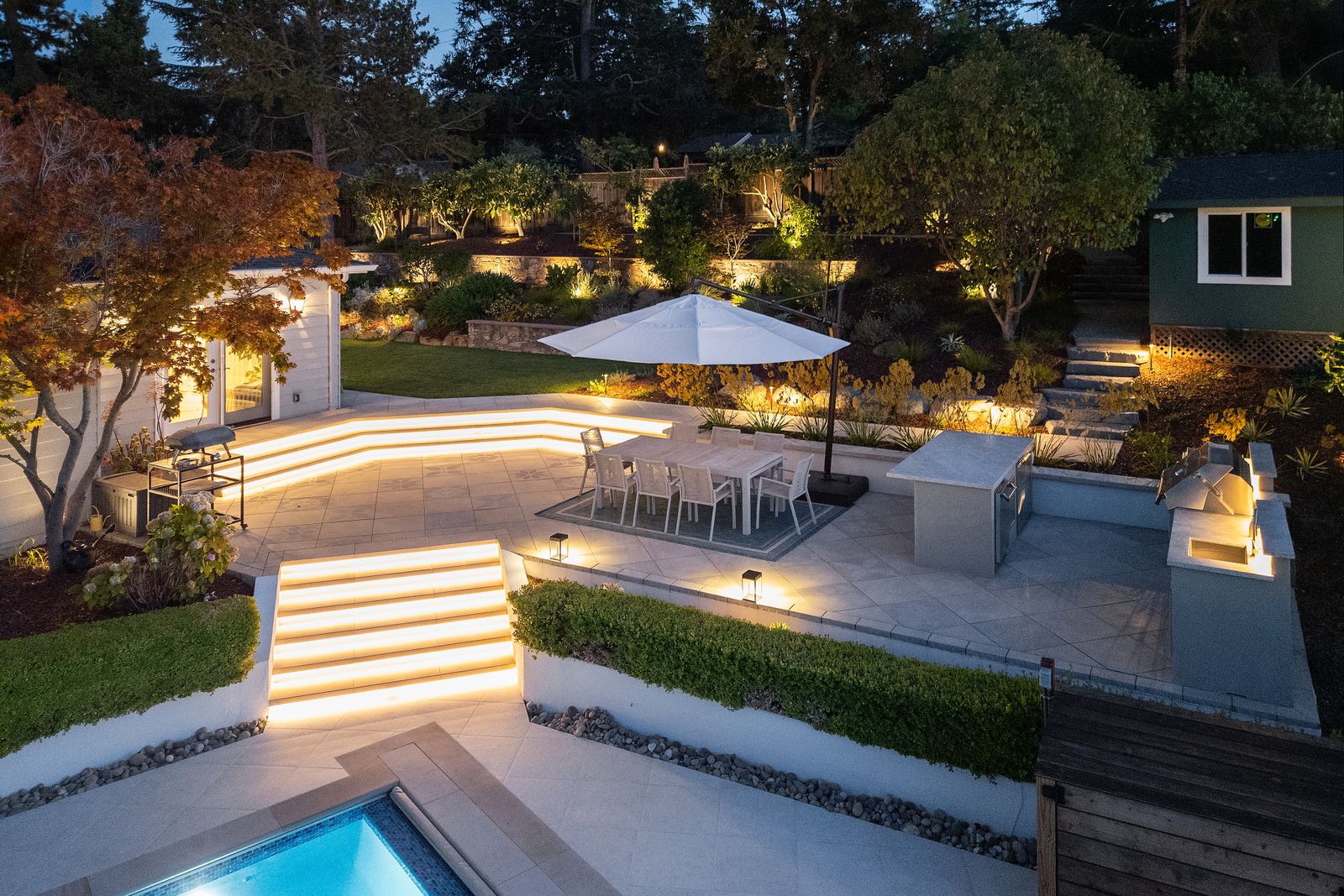 Twilight aerial of the terraced garden, pool lit below the stone retaining walls with the steps and terraces uplit