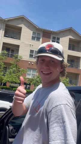 Smiling man in a ball cap giving a thumbs up outside an apartment building