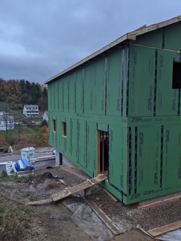 Green-sheathed house frame seen from the side, with a porch footing in the foreground