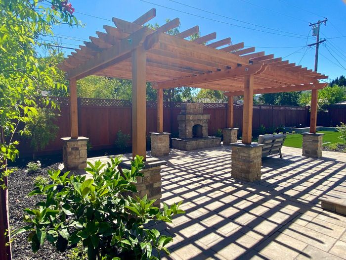 Wood pergola over a paver patio with a stacked stone fireplace in the far corner, stone-clad posts, green shrubs and dappled sunlight.