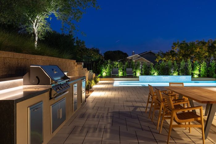Night view of a built-in outdoor kitchen with a stainless grill and dark stone counter beside a stepped retaining wall, a paver patio and a wooden dining table under a deep blue sky