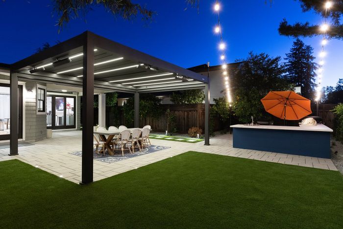 Dusk view across the turf to a louvered pergola with glowing strip lights over a dining table, and a blue kitchen island with an orange umbrella