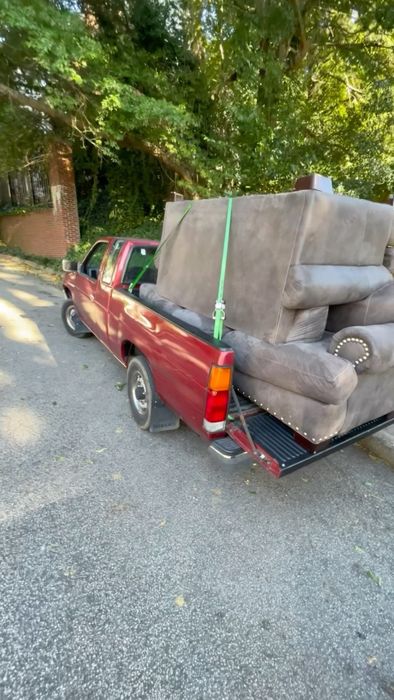 A large sectional couch strapped down on the back of a red pickup truck parked on a tree-lined street