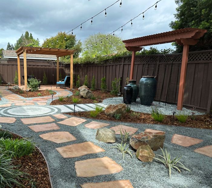 Terracotta flagstone stepping stones set in pale gravel past boulders and grasses, with two wood pergolas, large teal ceramic jars and string lights across the yard.
