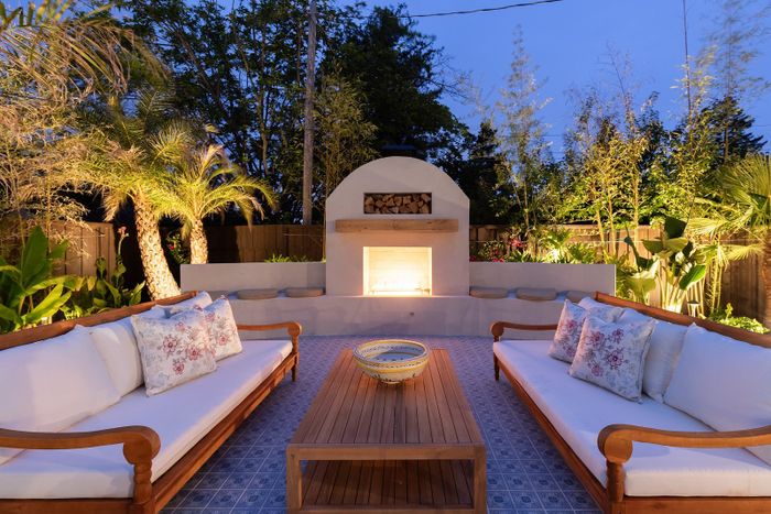 Glowing stucco fireplace with a stacked wood niche at twilight, two cushioned teak daybeds on a blue patterned tile floor and uplit palms and bamboo