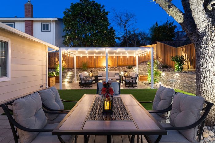 Dusk view across a dining table to a white louvered pergola with string lights over lounge seating, a stacked-stone wall and wood fence lit behind