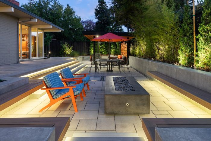Twilight view of a paver terrace with a concrete fire table, striped lounge chairs, lit steps and a wood-roofed pavilion with an orange umbrella