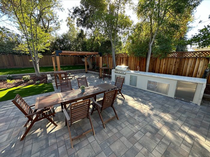 Paver patio with a wood dining table and chairs, a long built-in kitchen island with a grill and refrigerator, a wood pergola with string lights and a green lawn behind.