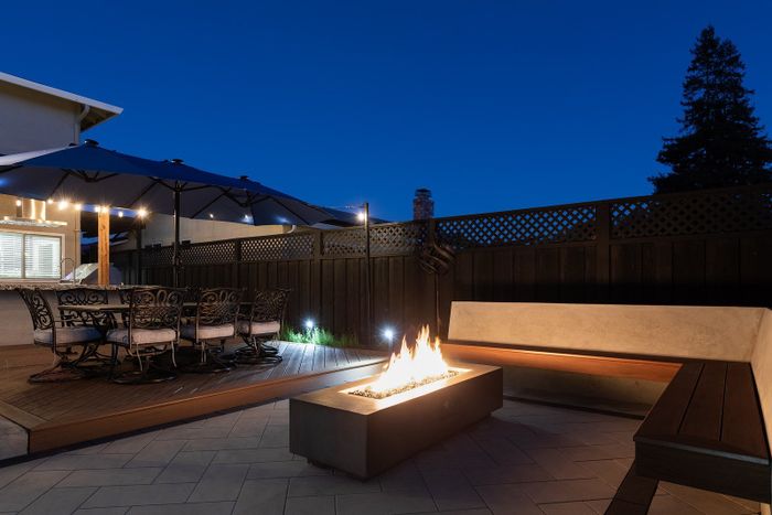 Twilight view across a pale paver court to a linear fire table and corner bench, with a dining table under a large umbrella on a wood-look deck and a covered kitchen beyond