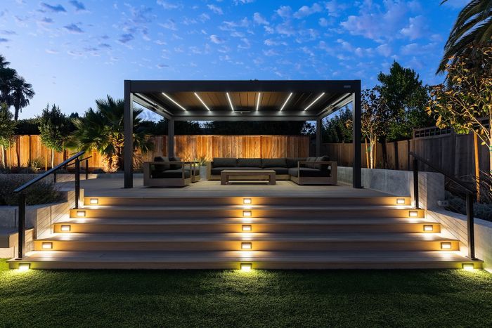 Twilight view of a raised deck with five wide lit steps, cable railings and a louvered pergola lit with white strip lights over a dark sofa lounge