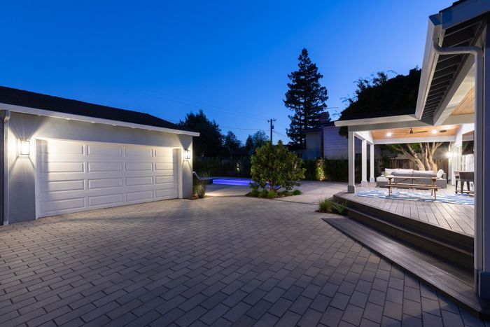 Blue-hour view of a paver driveway court in front of a white garage, with a covered deck and lounge seating glowing at the right
