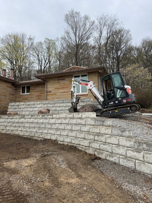 Compact excavator working above a long stacked block wall beside a brick house