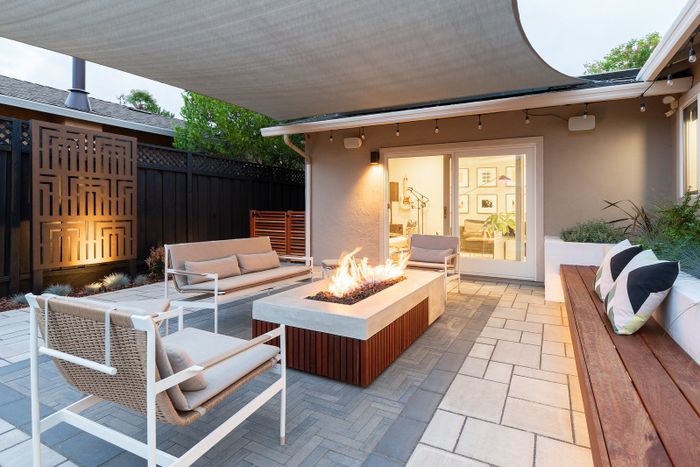 Gas fire table with an ipe base and built-in bench on a herringbone paver patio at dusk, with a lit decorative screen and shade sail overhead
