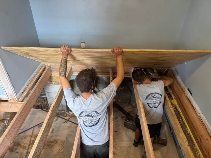 Two crew members lifting a sheet of subfloor over new floor joists