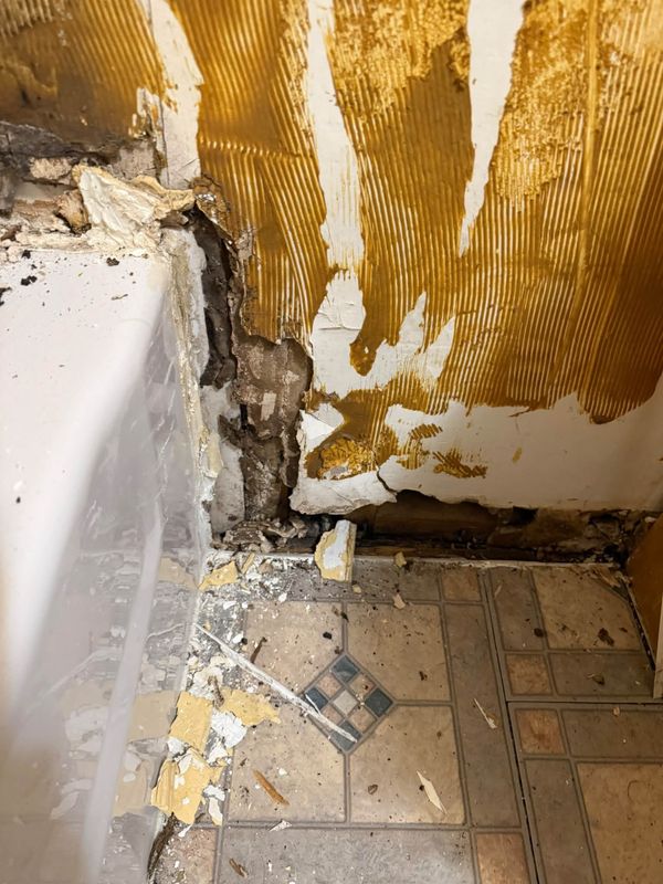 The corner beside a bathtub with crumbled wall board and debris on the vinyl floor