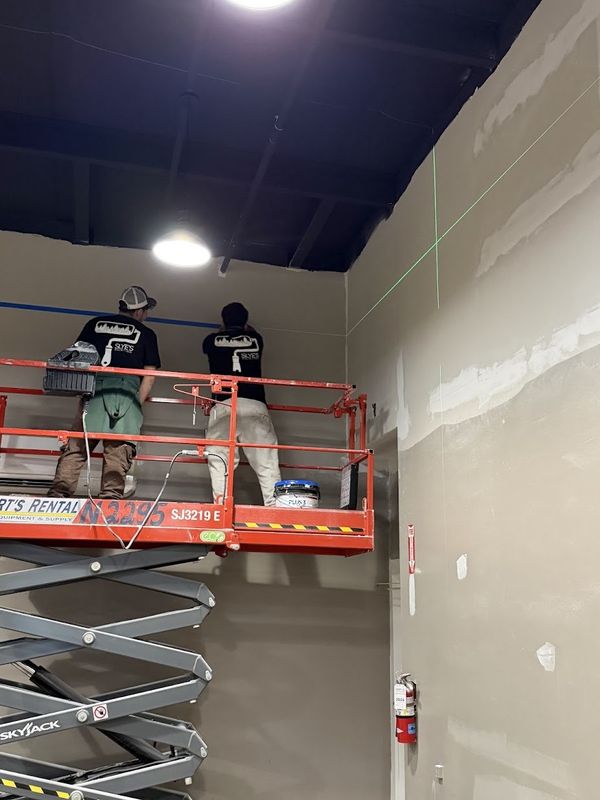Two Slye's Painting crew members on a scissor lift painting new drywall walls under a dark ceiling