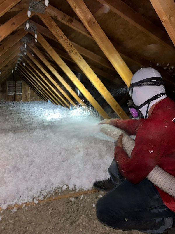 Foam Pro crew member in a red shirt and respirator blowing white fiberglass insulation across a Phoenix attic
