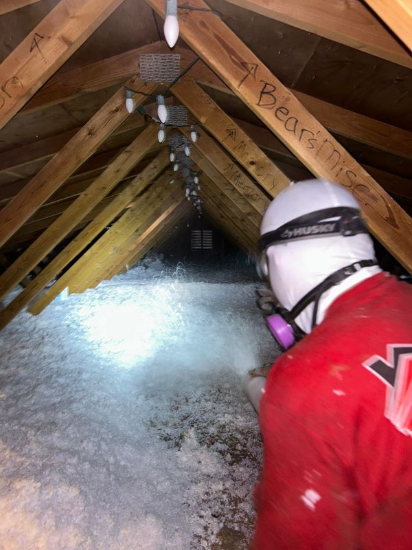 Foam Pro technician in a red shirt and respirator blowing white fiberglass insulation across an attic