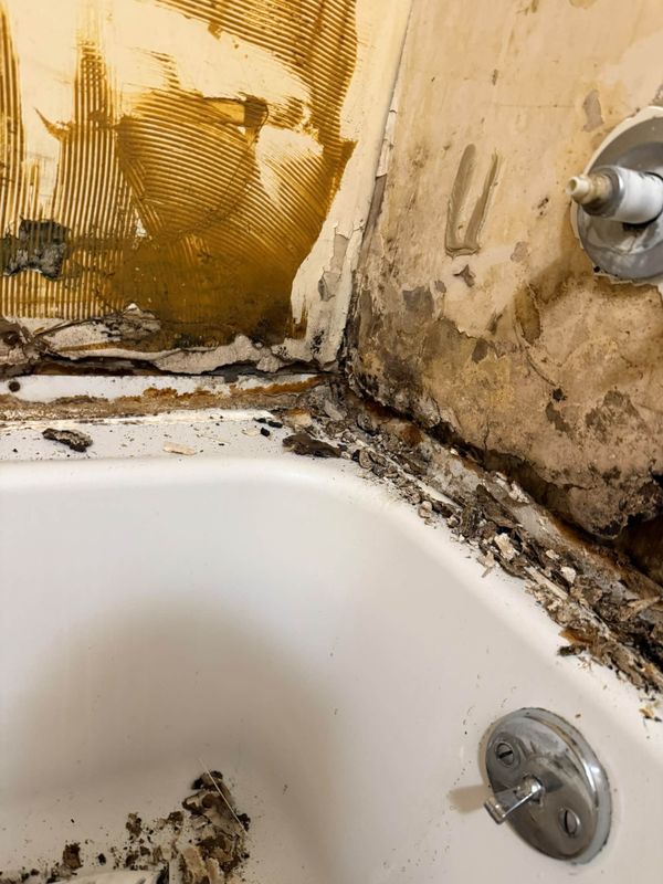 Water damaged wall stripped back above a bathtub, with debris collected in the corner