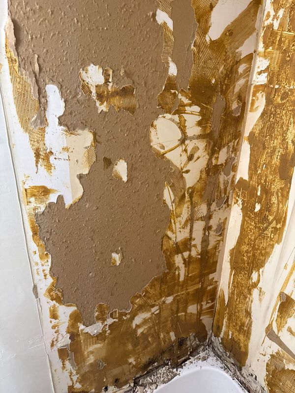 A bathroom wall with old textured paint peeling away from adhesive-covered board above a tub