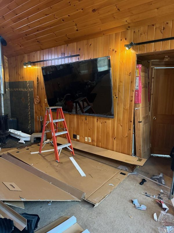 Large television mounted on a wood plank wall with a stepladder and packaging on the floor below